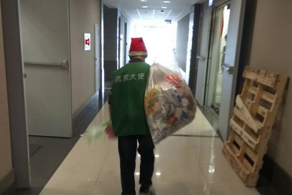 一位身穿綠色背心、頭載聖誕帽的清潔人員手揹垃圾袋的背影。